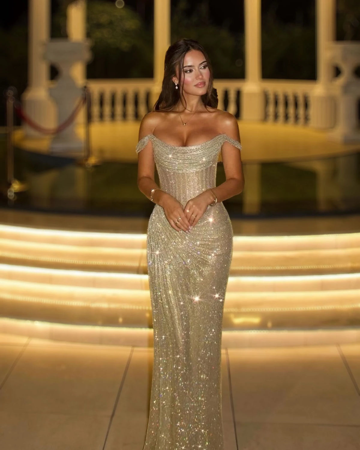 Full-length champagne crystal corset evening gown with draped off-shoulder neckline on illuminated steps