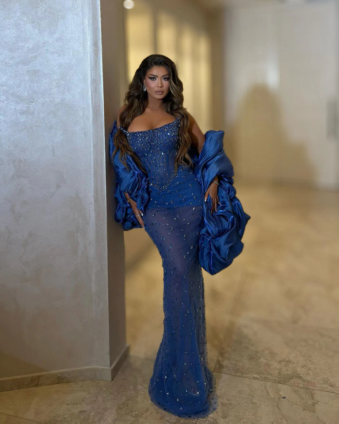 Front view of sapphire blue crystal mermaid evening gown with fitted waist and sculptural voluminous satin drapes framing the arms
